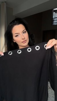 Črna oblačila s grometnimi detajli ob izrezu, gladko teksturo in ohlapnim krojem. Poudarjena v rokah za podroben pogled.