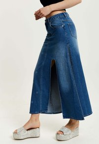 Denim maxi skirt with a front slit, blue hue, straight shape, and stitching details. Paired with silver platform sandals.