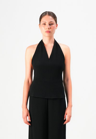 Femme avec les cheveux tirés en arrière, portant un top noir sans manches et un pantalon noir, se tenant devant un fond blanc.