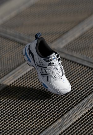 Witte en zwarte sportschoen met blauwe accenten, zwevend boven een getextureerde metalen roostervloer in natuurlijk licht.