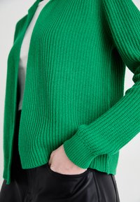 Cardigan tricoté vert avec une texture côtelée, à l'ouverture frontale et aux manches longues. Associé à un pantalon noir ajusté.