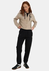 Felpa con cappuccio beige con zip, dettagli neri e coulisse, abbinata a pantaloni della tuta neri. Le scarpe da ginnastica presentano colori nero, bianco e marrone.