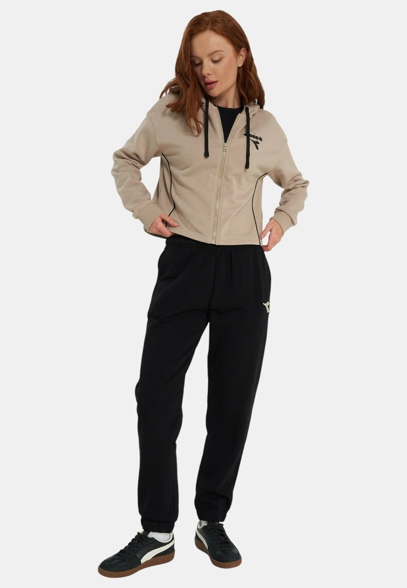 Felpa con cappuccio beige con zip, dettagli neri e coulisse, abbinata a pantaloni della tuta neri. Le scarpe da ginnastica presentano colori nero, bianco e marrone.
