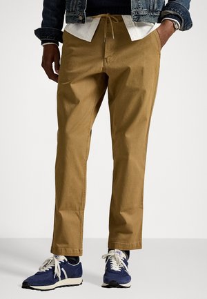 Tan bomullsbukser med rett benfôr, med elastisk strikk i livet. Kombinert med blå joggesko og en denimjakke.