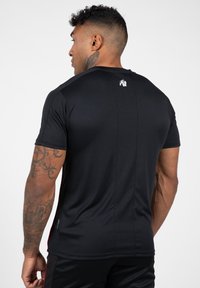 Zwarte sportieve T-shirt met korte mouwen, een gladde textuur, met een subtiele logo op de rug. De stof is rekbaar en aansluitend.