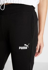 Schwarze Baumwoll-Sweatpants mit geripptem Bund, seitlichen Taschen und einem weißen Puma-Logo auf dem linken Oberschenkel. Glatte Textur.