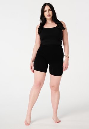 Un haut sans manches noir associé à un short noir taille haute, tous deux fabriqués dans un tissu lisse et extensible. Le mannequin se tient pieds nus sur un fond blanc.