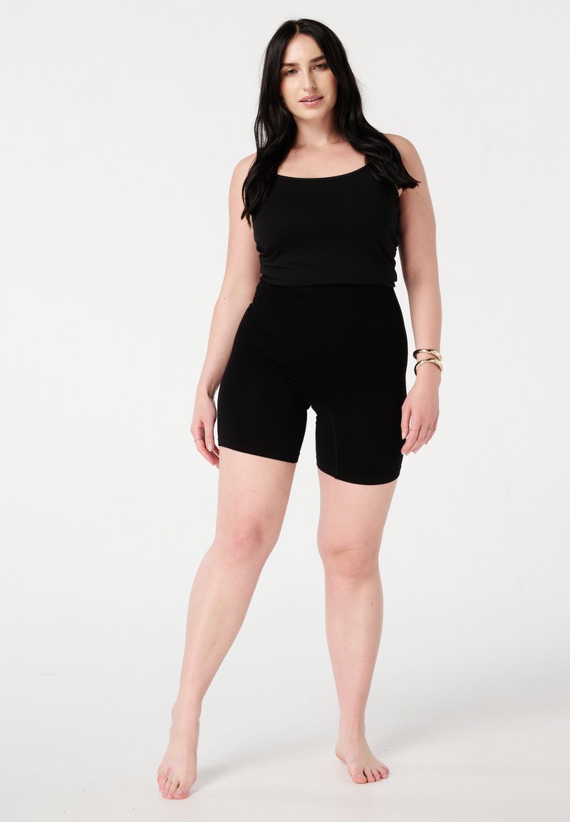Un haut sans manches noir associé à un short noir taille haute, tous deux fabriqués dans un tissu lisse et extensible. Le mannequin se tient pieds nus sur un fond blanc.