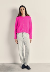 Moteris stovi prie smėlinės sienos, dėvi ryškiai rožinį užsegamą megztinį, šviesiai mėlynas džinsines kelnes ir juodus lakuotus batelius, rankos atsipalaidavusios šonuose.