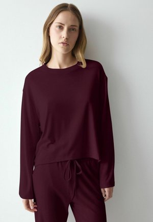 Young woman with light hair wearing a dark burgundy long-sleeve top and matching pants standing against a white wall.