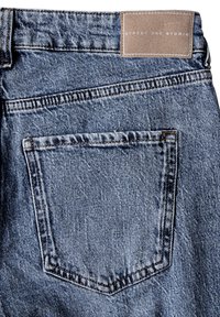 Jeans de denim en azul claro con un acabado texturizado, que presentan un bolsillo trasero y una etiqueta de marca de cuero marrón en la cintura.