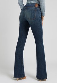 Pantalones vaqueros acampanados de color azul oscuro, hechos de mezclilla, con dos bolsillos traseros y una etiqueta de cuero en la cintura. Textura suave, diseño clásico.