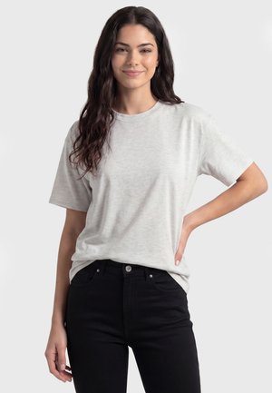 Młoda kobieta z długimi ciemnymi włosami uśmiecha się, ubrana w jasnoszary t-shirt z krótkim rękawem i czarne spodnie, stoi z jedną ręką na biodrze.