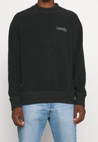 Uomo che indossa un felpa nera Levi's con un piccolo logo bianco sul petto e jeans blu chiaro, in piedi contro uno sfondo bianco.