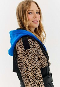 Sako z leopardím potlačou s modrou kapucňou, obsahujúce čierne akcenty a štruktúrovaný dizajn. Hladká textúra a priliehavý strih.