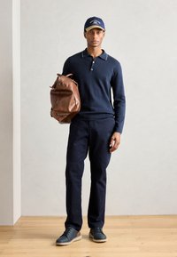 Polo de manga larga azul marino con un cuello en contraste, pantalones oscuros, bolsa de cuero marrón y zapatillas azules sobre un suelo de madera.