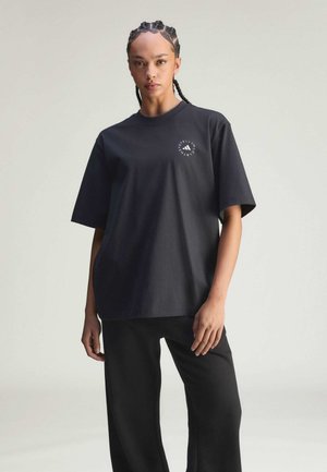 Jeune personne aux cheveux tressés portant un t-shirt noir oversize avec un logo blanc circulaire sur la poitrine et un pantalon noir assorti.