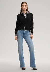 Μαύρη ζακέτα με κουμπιά, φορεμένη πάνω από πουκάμισο με λεοπάρ σχέδιο, σε συνδυασμό με ανοιχτό μπλε τζιν καμπάνα και μαύρα παπούτσια με τακούνι.