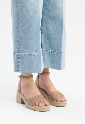 Sandali con tacco in suede beige, dotati di un ampia fascia anteriore, cinturino alla caviglia e suola spessa. Indossati con jeans a gamba larga di colore blu chiaro.