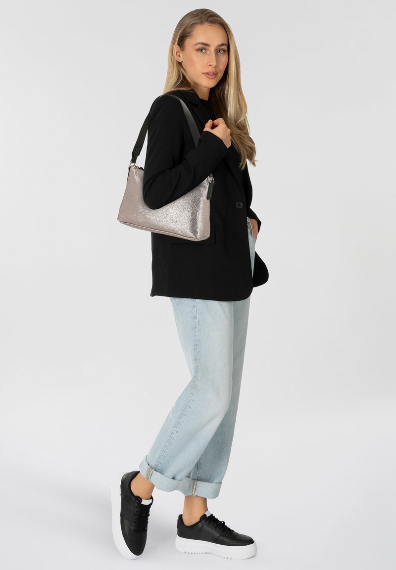 Metalen zilveren handtas met een gestructureerde vorm en een verstelbare zwarte schouderband, gedragen over een zwarte blazer, in combinatie met lichtgekleurde jeans en zwarte sneakers.