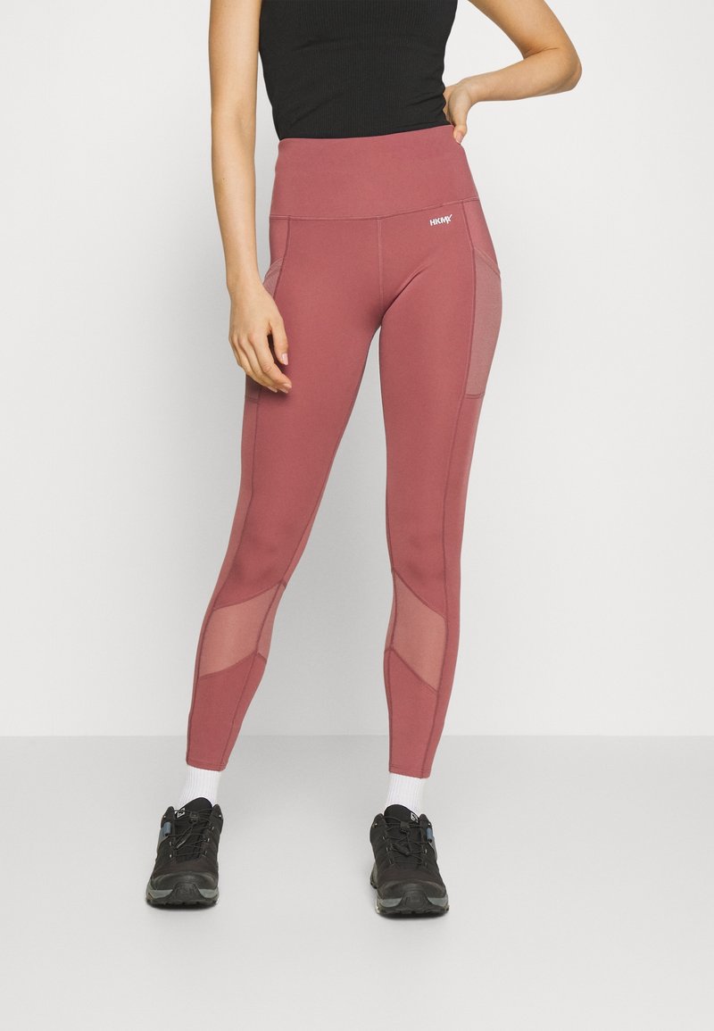 Höga leggings i dämpad ros med meshpaneler på sidorna, tillverkade av stretchigt tyg, tillsammans med svarta träningsskor.