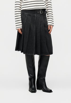 Czarna, plisowana spódnica o długości do połowy łydki z boczną klamrą, noszona z czarnymi, sięgającymi kolan skórzanymi kozakami oraz białą, wzorzystą bluzką z długim rękawem.