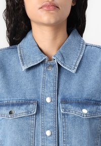 Ανοιχτό μπλε τζάκετ από τζιν με κλασικό γιακά, μεταλλικά κουμπώματα και δύο τσέπες στο στήθος με ασημένιες λεπτομέρειες. Ορατό υφασμάτινο σχέδιο.