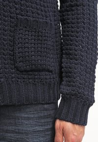 Tmavě modrý pletený svetr s texturovaným vzorem a malou přední kapsou, nošený s tmavě modrými džíny, ukazující manžetu a ruku.