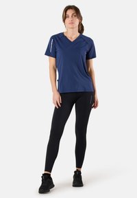 Marineblauw sporttops met korte mouwen en V-hals, met witte accentlijnen aan de zijkant, gecombineerd met zwarte lange leggings met een zijzak. Zwarte sneakers.