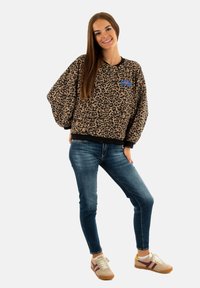 Sweatshirt con stampa leopardata e rifiniture nere, vestibilità ampia, con una toppa blu sul petto, abbinato a jeans blu e sneakers beige con strisce color bordeaux.