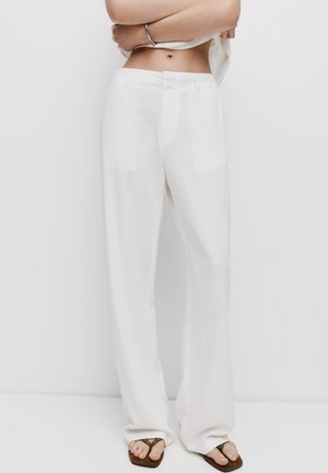 Pantaloni larghi bianchi in lino con una texture pulita e liscia; presentano tasche frontali e una vestibilità dritta, abbinati a sandali di colore scuro.