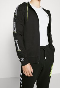 Uomo in felpa nera con zip, con nastro logo bianco e giallo sulle maniche, t-shirt bianca e pantaloni neri con patch logo giallo fluo.