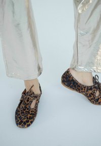 Scarpe in tessuto leopardato con design a taglio e dettaglio a fiocco, abbinate a pantaloni metallici argento lucido. Suole piatte.