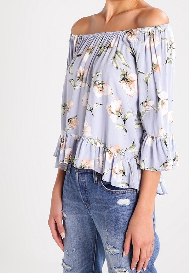 Femme portant un chemisier floral violet clair à épaules dénudées avec des manches volantées et un jean bleu délavé.