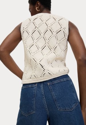 Trui zonder mouwen in crème met een open ruitpatroon. De achterkant heeft een afgeronde zoom en is gecombineerd met blauwe denim high-waisted jeans.