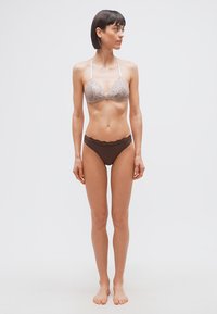 Γυναίκα που στέκεται ξυπόλυτη, φορώντας ένα ανοιχτό μπεζ δαντελένιο μπουστάκι και σκούρο καφέ εσώρουχο με δαντέλα στα τελειώματα, κοιτάζοντας προς τα δεξιά της μπροστά σε απλό λευκό φόντο.