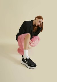 Kvinde med langt rødt hår, der croucher, iført en sort blazer, lyserød plisseret nederdel, hvide sokker og sorte sneakers med tykke mønstrede såler.