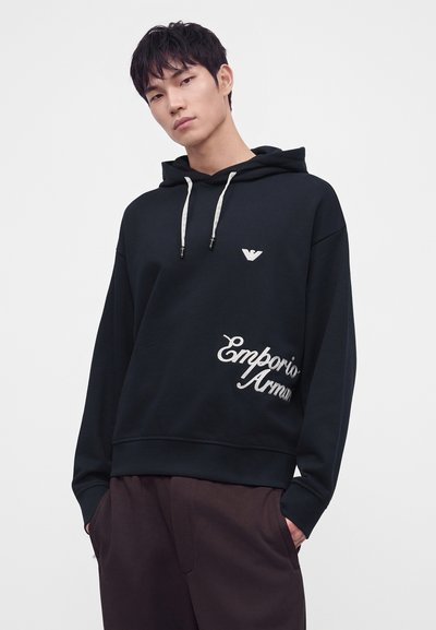 Jeune homme portant un sweat à capuche noir Emporio Armani avec logo blanc et pantalon foncé, debout devant un fond blanc uni.