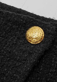 Zwart, gestructureerd stof met een opvallende gouden knop, die een gecanneleerd ontwerp en lauwerkransdetails heeft, wat het algehele uiterlijk van het kledingstuk verbetert.