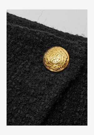 Czarna, teksturowana tkanina z wyraźnym złotym guzikiem, ozdobiona wytłaczanym wzorem i detalami w kształcie wieńca, podkreślającymi ogólny wygląd odzieży.