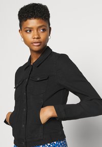 Giubbotto di jeans nero con colletto, chiusura a bottoni vorne e due tasche sul petto. Presenta maniche lunghe e una silhouette aderente.
