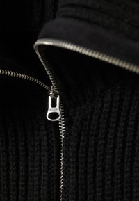 Schwarze gestrickte Jacke mit Reißverschluss verfügt über einen silbernen Reißverschluss und ein Etikett mit der Aufschrift "WEEKDAY." Strukturierter Stoff mit vertikalem Rippdesign.