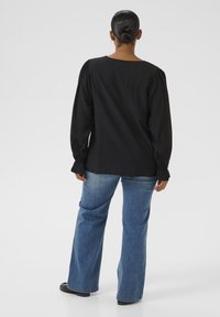 Schwarzes Langarmshirt mit gerafften Ärmeln, kombiniert mit hellblauen, weit geschnittenen Denim-Jeans. Der Stoff wirkt weich und leicht strukturiert.