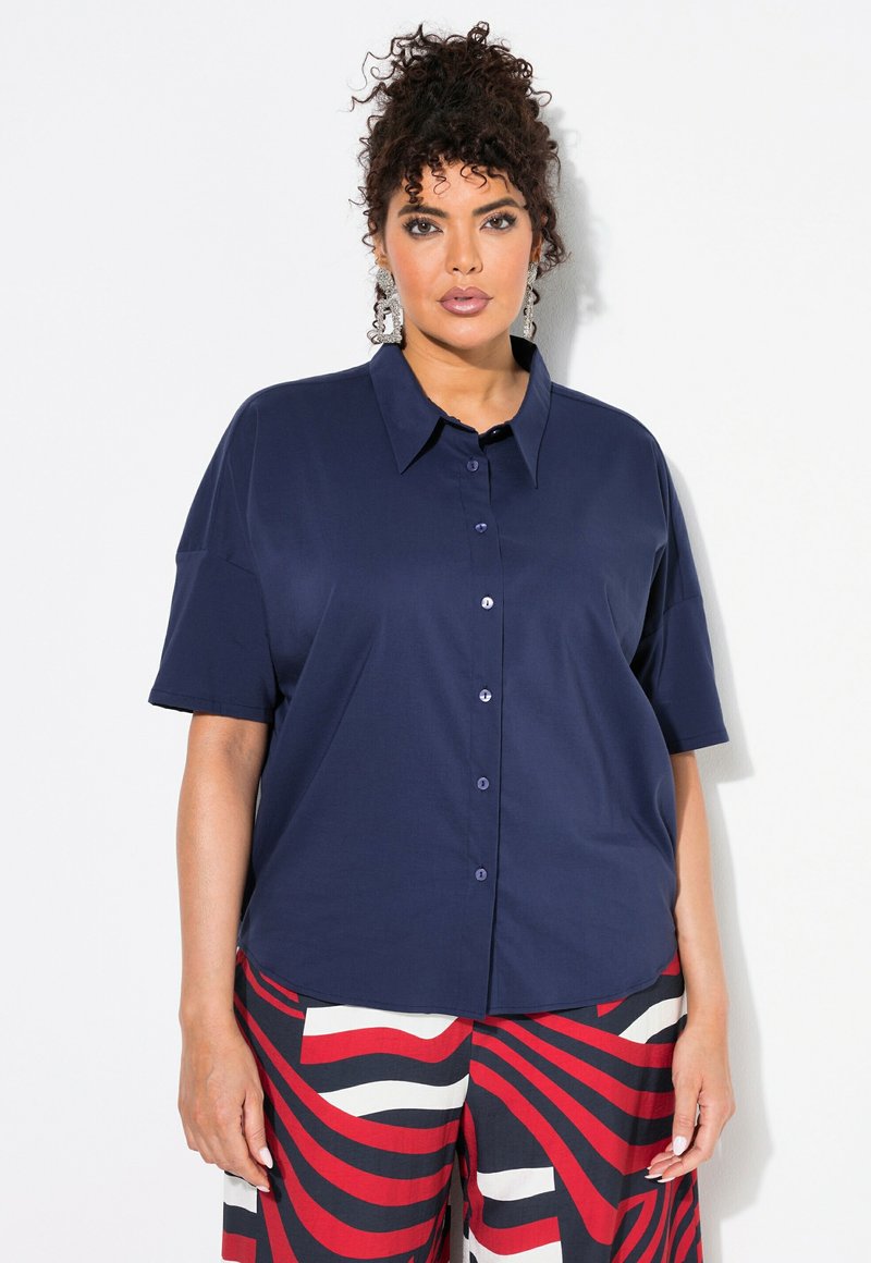 Camicia corta blu navy con colletto a punta e chiusura con bottoni. Tessuto liscio con una vestibilità rilassata, abbinata a pantaloni a motivi rossi e bianchi.