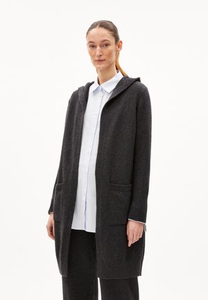 Dunkelgrauer, langer Cardigan mit Kapuze aus weichem Material. Verfügt über zwei frontale Taschen und gerippte Bündchen. Über einem hellblauen Hemd getragen.