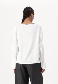 Blusa bianca con polsini arricciati, tessuto leggero, vestibilità rilassata, scollatura rotonda. Abbinata a pantaloni grigi scuri su misura.