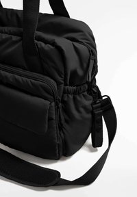 Črna oblazinjena torbica s predalom z zadrgo, nastavljivim trakom ter teksturirano tkanino, opremljena s trdnimi ročaji in kovinskimi dodatki.