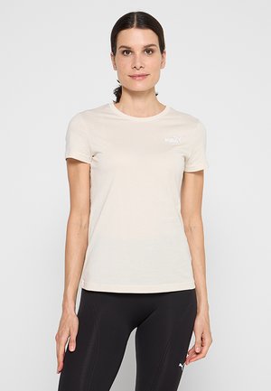 Mujer con pelo oscuro recogido, vistiendo una camiseta Puma beige claro y mallas negras Puma, de pie frente a un fondo blanco liso.