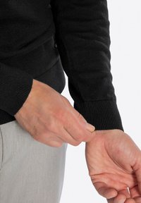 Czarny dzianinowy sweter z prążkowanymi mankietami, na którym widać rękę poprawiającą rękaw. Materiał wydaje się miękki z lekko teksturowanym wykończeniem.