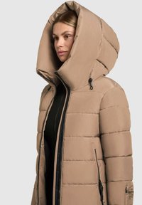 Beige pufferjakke med en stor, struktureret hætte. Har lynlåslukning, tekstureret stof og diskret branding på ærmet.